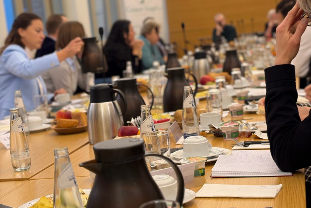 Langer Frühstückstisch mit Kaffee, Obst und Unterlagen beim parlamentarischen Frühstück im Bundestag, Teilnehmende im Gespräch über sicherheitspolitische Fragen.